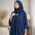 A woman wearing a navy blue modest outfit consisting of a long abaya dress and matching hijab, standing in a softly lit living room with a sofa and lamp in the background.