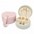 A round jewelry box shown open and closed on a white background. The closed box is light pink with a strap and snap closure.